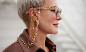 Kobieta z krótkimi blond włosami nosi okulary z ozdobnym złotym łańcuszkiem i wiszące złote kolczyki z perełkami.
