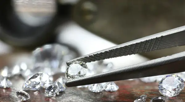Szczypce trzymają błyszczący diament, obok leżą inne kamienie. To nie jest kaboszon, lecz brylant.